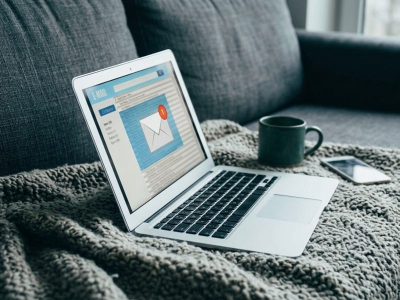 Open laptop showing an email inbox on a grey blanket beside a mug