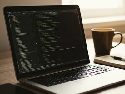 Laptop showing colorful programming code next to a mug of coffee and a notebook