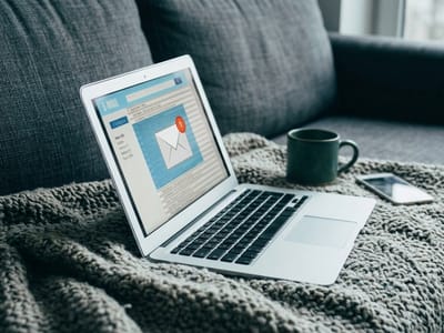 Open laptop showing an email inbox on a grey blanket beside a mug